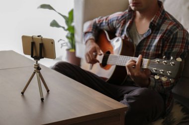 Ekoseli ekose gömlekli müzik koleji öğrencisi akustik gitar egzersizi yapıyor, dizüstü bilgisayardan notlar okuyor. Karantina sırasında evde müzik dersi alan bir adam. Arkaplan, kapat