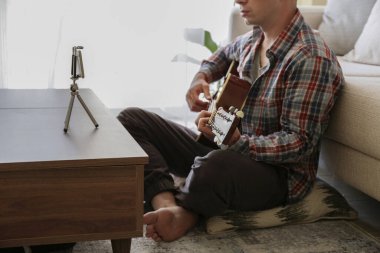 Ekoseli ekose gömlekli müzik koleji öğrencisi akustik gitar egzersizi yapıyor, dizüstü bilgisayardan notlar okuyor. Karantina sırasında evde müzik dersi alan bir adam. Arkaplan, kapat