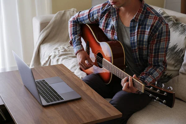 Music college hipster student in checkered plaid shirt practicing acoustic guitar exercise, reading notes from laptop computer. Man taking an online musical courses at home during quarantine. Background, close up