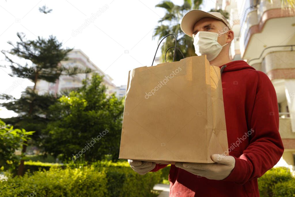 Delivery guy wearing red hoodie, disposable gloves and mask for coronavirus protection, holding blank brown paper grocery bag outside of high rise apartment building. Close up, copy space.