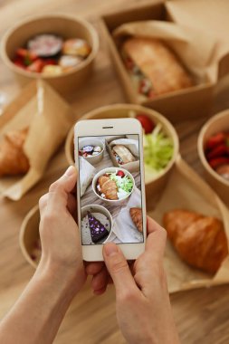 Kadın yemek blogcusu dışarıdan gelen yemekleri inceliyor, telefonda video çekiyor. İtalyan sandviçi, Fransız kruvasanı w / somon, çikolatalı cheesecake. Kapat, üst görünüm, pov, kopyalama alanı, ahşap masa arkaplanı.