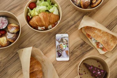 Ahşap mutfak masasında farklı servis yemekleri. İtalyan panini sandviçi, somonlu Fransız kruvasanı, krep, çikolatalı cheesecake, telefon ve yemek fotoğrafı. Kapat, üst görünüm, kopyalama alanı, arkaplan.