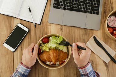 Yemek arası konsepti. İş yerinde dışarıdan yemek yiyen bir adamın görüntüsü. Laptop, boş ekran telefon ve not defteri. Fransız kruvasanı ve salata. Üst görünüm, kapat, kopyala alanı, tahta masa arkaplanı