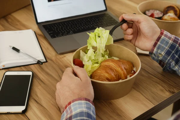 Lunch break concept. POV shot of man eating takeout food by his workspace. Laptop, blank screen phone & notebook. French croissant with salad. Top view, close up, copy space, wooden table background