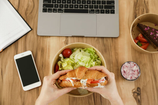 Концепция перерыва на обед. Женщина ест еду на вынос на рабочем месте. Ноутбук, пустой экран телефона и ноутбука. Французский круассан, салат, шоколадный торт. Вид сверху, POV, крупный план, копировальное пространство, деревянный фон
