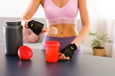 Evde eğitim konseptinde kal. Sportif vücutlu çekici bir kadın egzersiz sonrası protein içeceği içiyor. Kadın spor kıyafetleri içinde güzel bir kadın elinde kırmızı bir tokalaşma aleti tutuyor. Kapat, arkaplanı kopyala