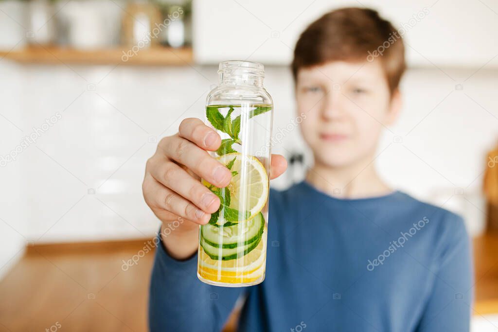 Teenager boy holding glass bottle infused detox water with cucumber, lemon and mint kitchen on background. Diet, healthy eating, weight loss concept. Copy space