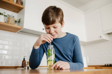Genç çocuk arka planda paslanmaz çelik saman mutfağı kullanarak salatalık, limon ve nane şekeri ile detoks suyu içiyor. Sağlıklı yaşam için sağlıklı bir içecek. Boşluğu kopyala