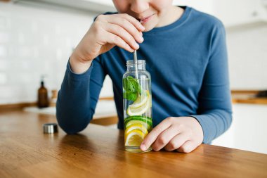 Genç çocuk arka planda paslanmaz çelik saman mutfağı kullanarak salatalık, limon ve nane şekeri ile detoks suyu içiyor. Sağlıklı yaşam için sağlıklı bir içecek. Boşluğu kopyala