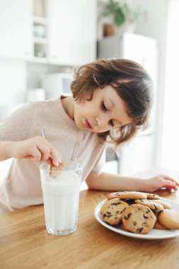 Küçük tatlı kız bardaktan çelik çubukla süt içiyor ve mutfak masasında oturmuş kurabiye yiyor. Çocuklarla evde plastik kullanımını azalt