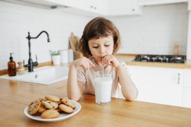 Küçük tatlı kız bardaktan çelik çubukla süt içiyor ve mutfak masasında oturmuş kurabiye yiyor. Çocuklarla evde plastik kullanımını azalt