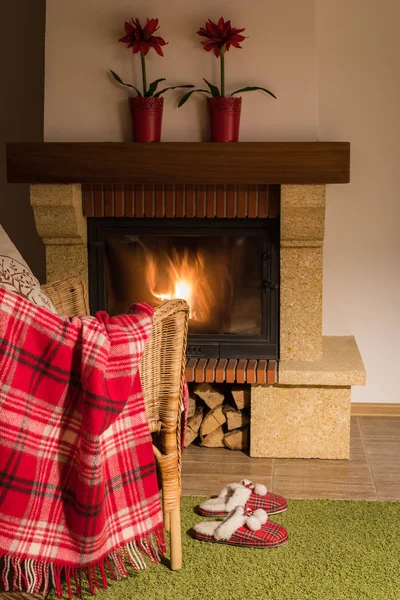 Wool warm blanket or plaid and red slippers with pompons on the background of burning fireplace