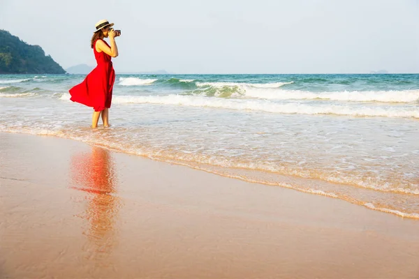 身穿红色衣服的女摄影师站在海滩上泡在水里 把照片拍摄到大海广阔而遥远的地方 蓝天背景和复制空间 现代女性生活方式的观念 — 图库照片
