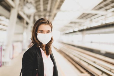Cute pretty Asian female in a black suit wearing medical hygiene protective mask standing alone waiting for train at station. Concept for businesspeople prevents health from coronavirus or COVID-19. clipart