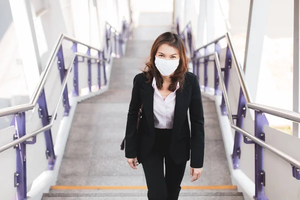 Beautiful Asian woman in a black suit wearing medical hygiene protective mask walking on stair in city to work at office. Idea for health care and prevent from risk of coronavirus epidemic outbreak.