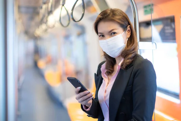 Bueatiful adult Asian businesswoman passenger commuter wearing medical hygiene protect mask. Standing and using cell phone inside empty subway or sky train car. Idea for abandoned city from COVID-19.