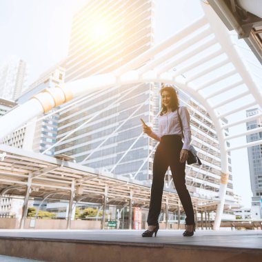 Yetişkin, güzel, orta yaşlı Asyalı bir ofis çalışanı. Elinde akıllı telefon olan ve iş binaları ve turuncu güneş ışığı arka plandaki cam duvara yansıyan büyük bir şehirde ofise yürüyen bir iş kadını..