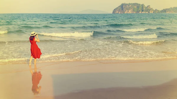身穿红色衣服的女摄影师站在海滩上泡在水里 把照片拍摄到大海广阔而遥远的地方 蓝天背景和复制空间 现代女性生活方式的观念 — 图库照片