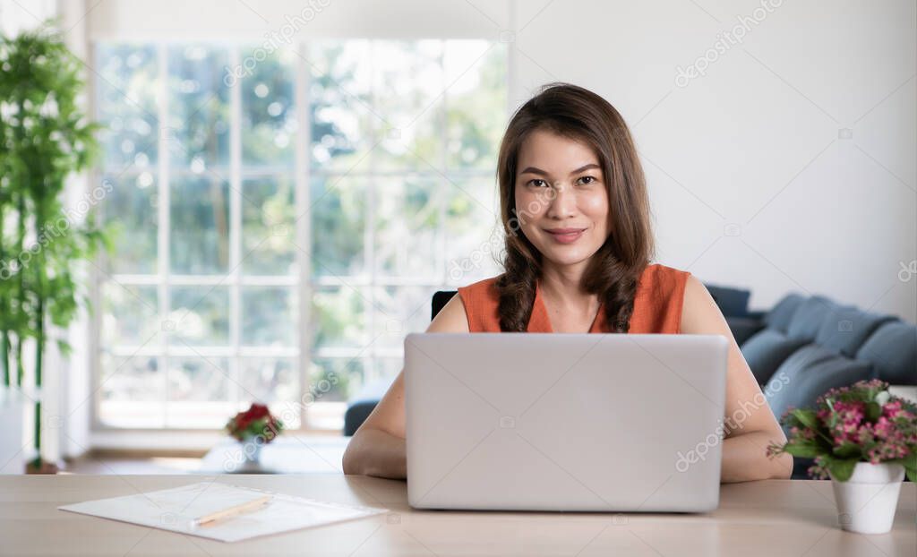 Beautiful middle age Asian businesswoman sitting at desk and using laptop notebook computer in apartment or condominium living room with easy and happiness. Idea for work at home or home base working.