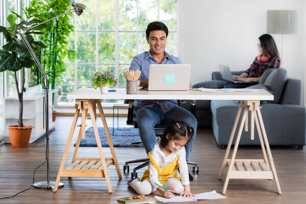 Mixed race family sharing time in living room. Caucasian father using notebook computer to work and half-Thai playing and painting under desk while Asian mother with laptop working her job on sofa.