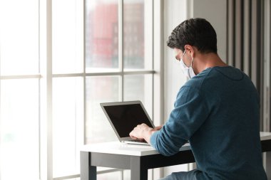 Tıbbi hijyen giyen beyaz bir adam maske takıyor. Oturma odasında dizüstü bilgisayar kullanıyor. Coronavirüs salgını sırasında sağlık hizmetleri karantinası ve evden çalışma fikri.