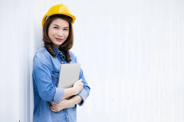 Beautiful Asian female engineer in jeans dress wearing yellow safety hard hat using laptop computer notebook doing job at construction site outside office. Idea for modern working woman.