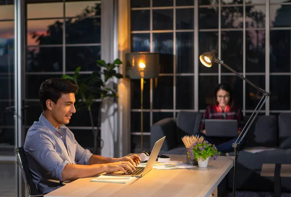 Caucasian businessman sitting happily on desk in living room under warm lamp and twilight blue sky outside house in background. Idea for working late and lifestyle of freelance, working at home couple