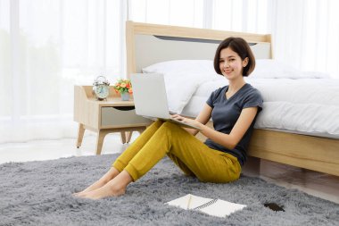 Genç, güzel, yarı Taylandlı bir kız yatak odasının yanında dizüstü bilgisayar kullanarak rahat bir evde kahve fincanıyla çalışıyor. Modern kadının ev yaşamından çalışma fikri..