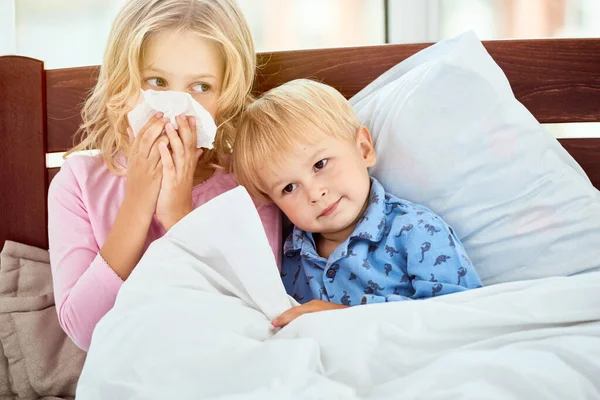 Just rest and sleep. Little sister and brother with runny noses suffering from flu or cold while lying in bed together at home. Virus disease