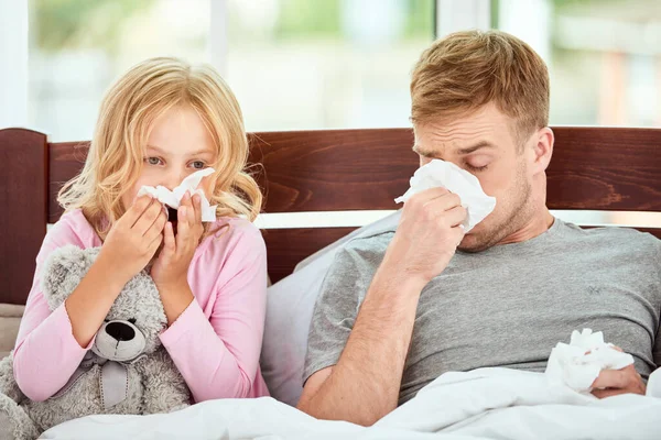 Family health. Young father and his daughter are suffering from flu or cold and runny nose while lying in bed together at home. Virus disease