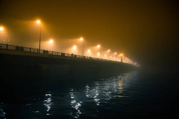 Una hermosa niebla nocturna sobre el río en el centro de la ciudad. Puente en la niebla, paisaje otoñal. Mirada suave, borrosa y brumosa. Colorido paisaje urbano místico de Riga, Letonia . — Foto de Stock