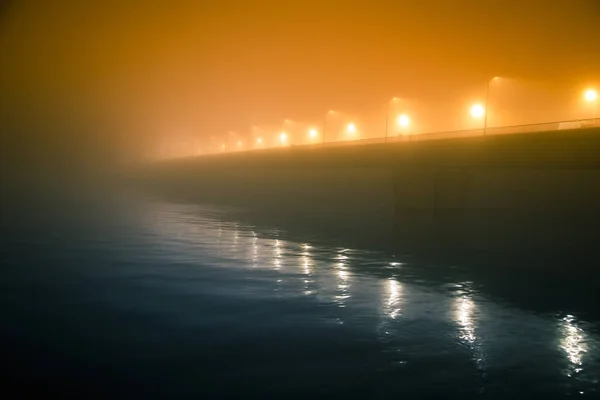 Una hermosa niebla nocturna sobre el río en el centro de la ciudad. Puente en la niebla, paisaje otoñal. Mirada suave, borrosa y brumosa. Colorido paisaje urbano místico de Riga, Letonia . — Foto de Stock