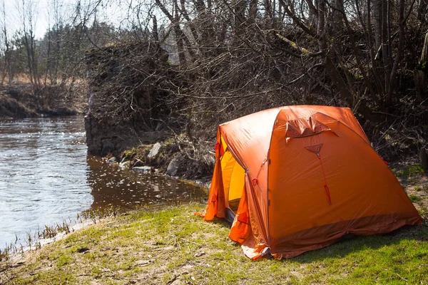 An orange tent built on the bank of the river. Hiking camp in the spring. Early spring scenery.