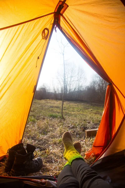 A person sitting in an orange tent, camp in the bank of the river in spring. Feet selfie of traveler. Relaxed, colorful look. Spring hiking.