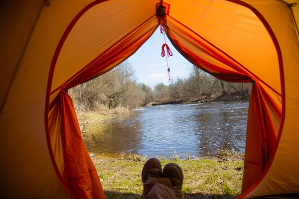 A person sitting in an orange tent, camp in the bank of the river in spring. Feet selfie of traveler. Relaxed, colorful look. Spring hiking.
