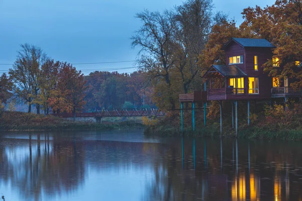 Retreat near lake in the evening. Wooden cottages on piles in oak forest. House with panoramic windows and lights on. Vacation getaway.