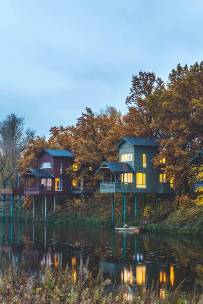 Retreat near lake in the evening. Wooden cottages on piles in oak forest. House with panoramic windows and lights on. Vacation getaway.