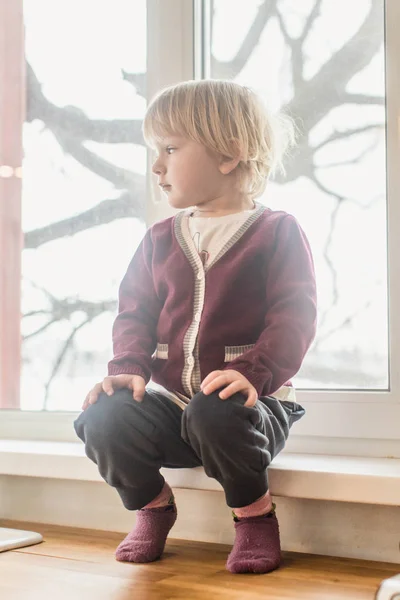 Cute adorable little boy in the kitchen sitting on wooden counter top, looking in the window.