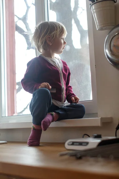 Cute adorable little boy in the kitchen sitting on wooden counter top, looking in the window.