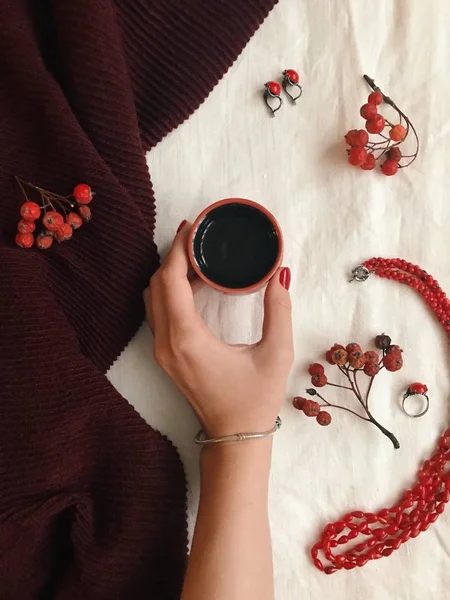 Винтажная модная квартира лежала с рукой держа чашку чая, женские аксессуары, шарф, красные бусины, серьги, кольцо и рябина, вид сверху — стоковое фото