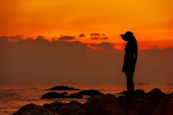 Silueta de una niña en un sombrero sobre el fondo de una puesta de sol de mar naranja . — Foto de Stock