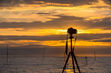 Bir deniz manzara parlak turuncu akşam gökyüzü arka plan üzerinde bir kamera ile üçayak silüeti.