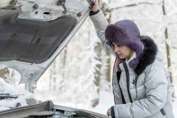 The girl lifted the hood from the car and looks at the engine.