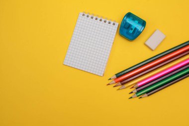 Renkli kalemler, silgi, silgi ve sarı arka planda beyaz bir not defteri, yazı için yer. Düz yatıyordu. Kırtasiye malzemeleri. okul malzemeleri