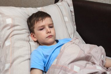 Çocuk hoşnutsuz bir yüzle yatakta yatıyor, hasta. Ev karantinası. Virüs. Grip. Yatak istirahatı