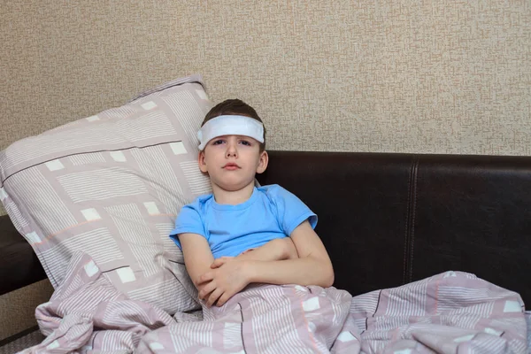 A sick child lies on a bed with a wet towel on his forehead. Headache in a child and high fever. Domestic quarantine. Virus