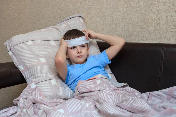 A sick child lies on a bed with a wet towel on his forehead. Headache in a child and high fever. Domestic quarantine. Virus