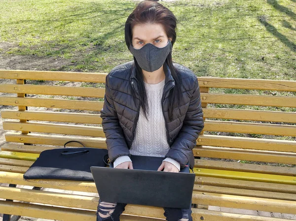 Self-isolation and work remotely during quarantine. A girl in a medical cloth bandage sits on a bench in a park and works on a laptop remotely.