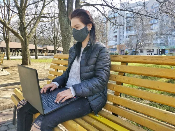 Self-isolation and work remotely during quarantine is an epidemic of coronavirus. A girl sits on a bench in a park and works on a laptop remotely. Portrait of a girl in a fabric mask