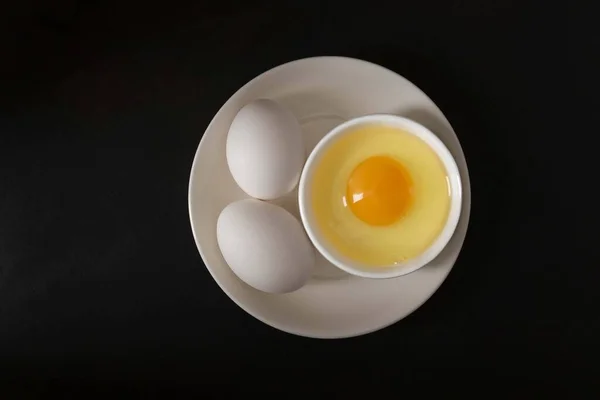 Aerial view of two white eggs: on a white plate two eggs, next to them on another plate, an egg white and an egg yolk, all on a black background